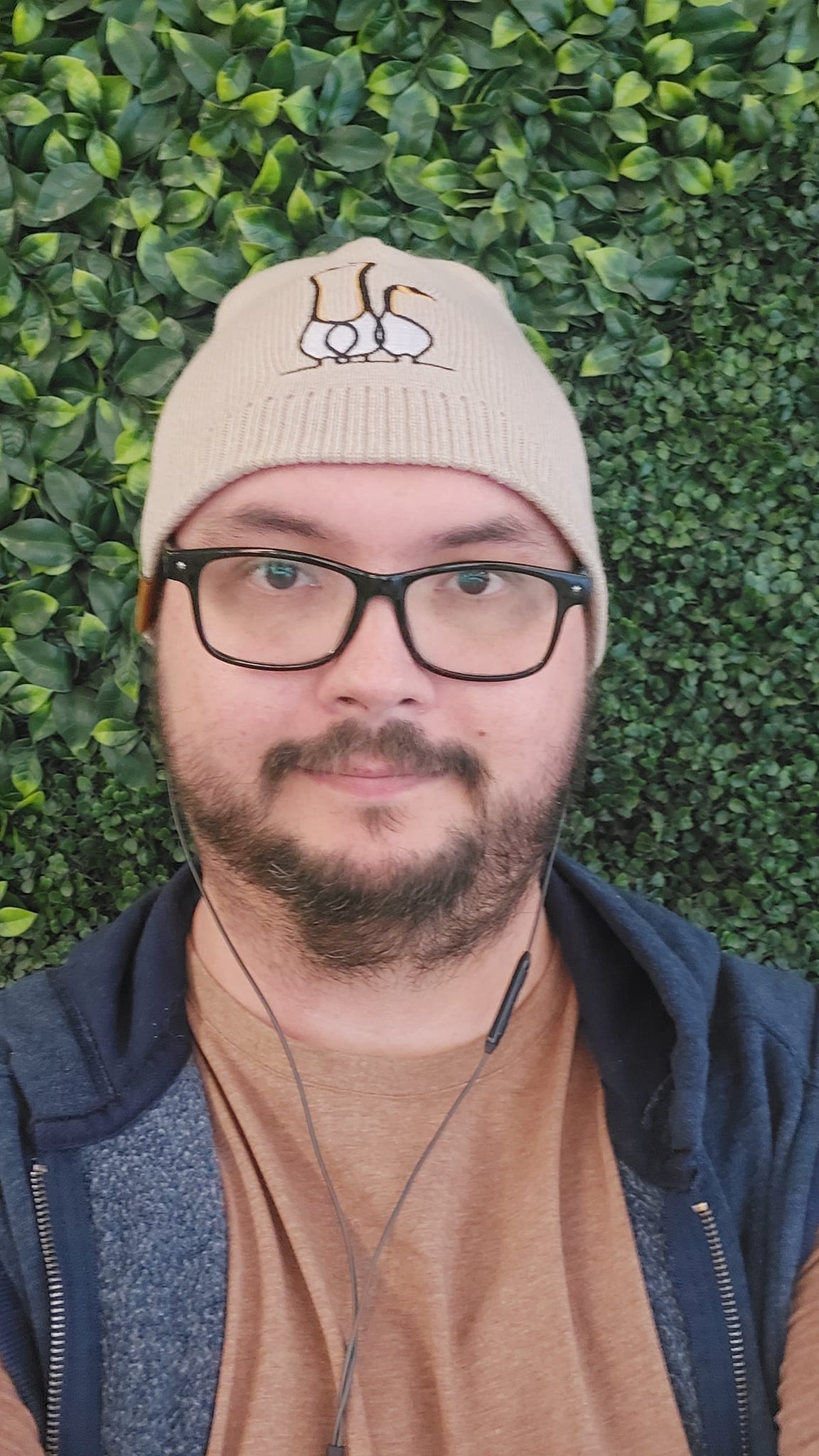 A man is dressed in street clothes standing in front of an ivy covered wall, he has a beard and moustache, a toque with geese on it, black rimmed glasses, a brown shirt and blue hooded sweater vest